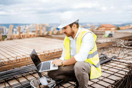 A construction engineer in a hard hat and safety vest works on a laptop.