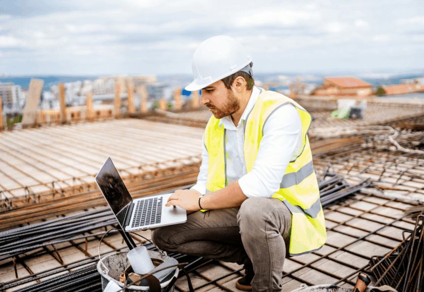 A construction engineer in a hard hat and safety vest works on a laptop.