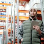 Warehouse worker in a beanie and vest holding a tablet in a storage aisle.