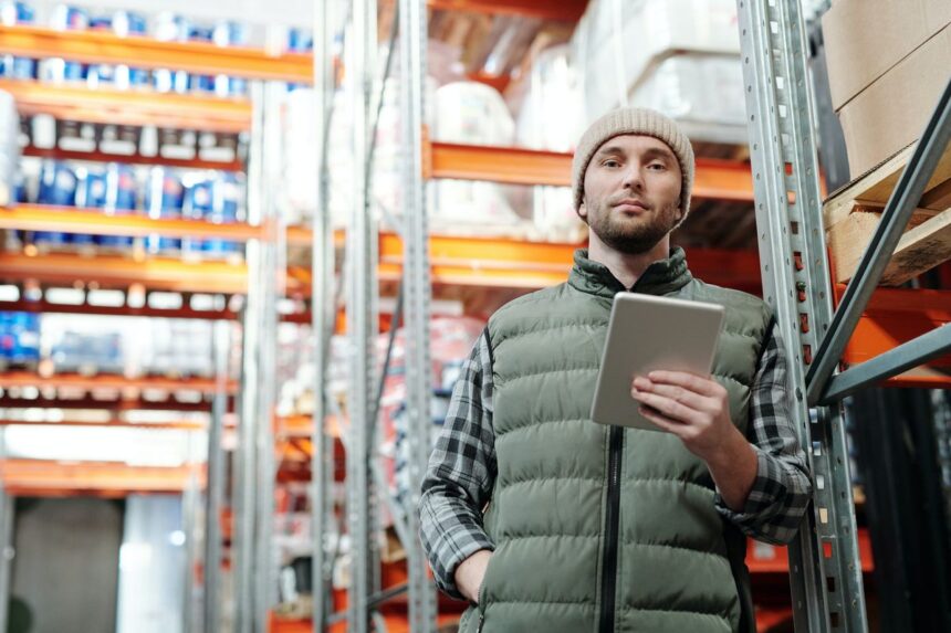Warehouse worker in a beanie and vest holding a tablet in a storage aisle.