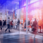 Blurry silhouettes of people walking past brightly lit, colorful store window displays.