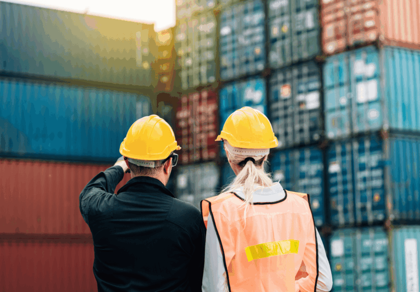 Two workers in hard hats standing before stacked shipping containers at a port.