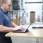 A warehouse worker stands at a desk typing on a laptop.