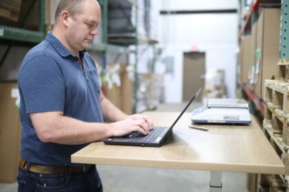 A warehouse worker stands at a desk typing on a laptop.
