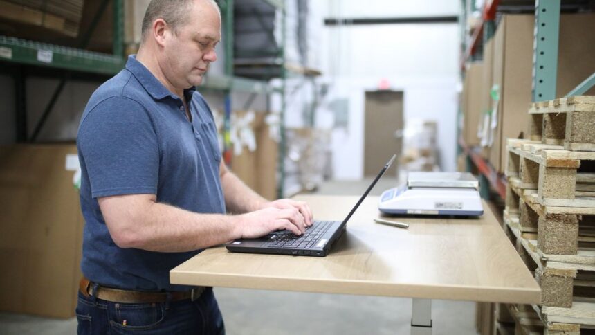 A warehouse worker stands at a desk typing on a laptop.