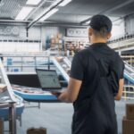 Warehouse worker holding a laptop with data, overlooking an automated package sorting conveyor belt.