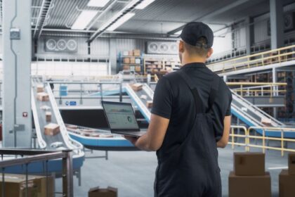 Warehouse worker holding a laptop with data, overlooking an automated package sorting conveyor belt.