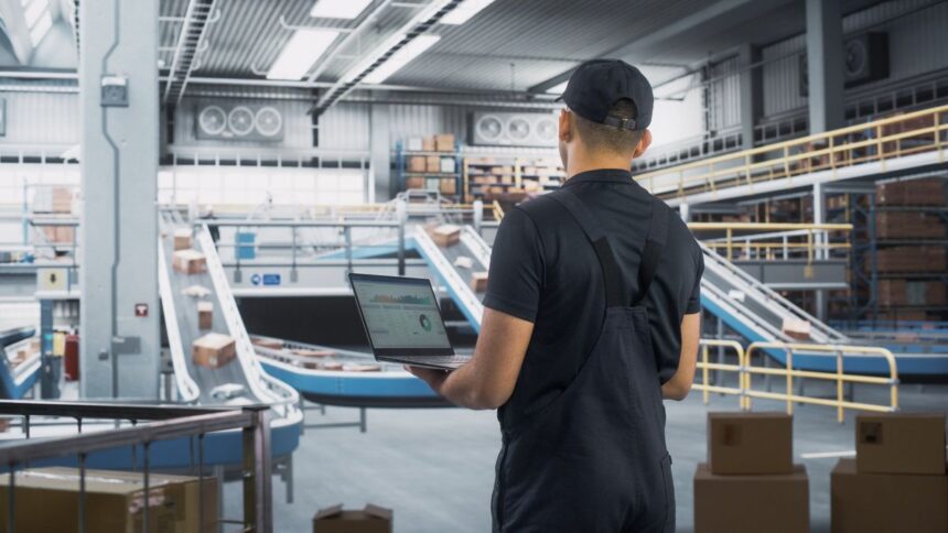 Warehouse worker holding a laptop with data, overlooking an automated package sorting conveyor belt.