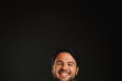 A smiling man with a beard wearing a green button-down shirt against a black background.