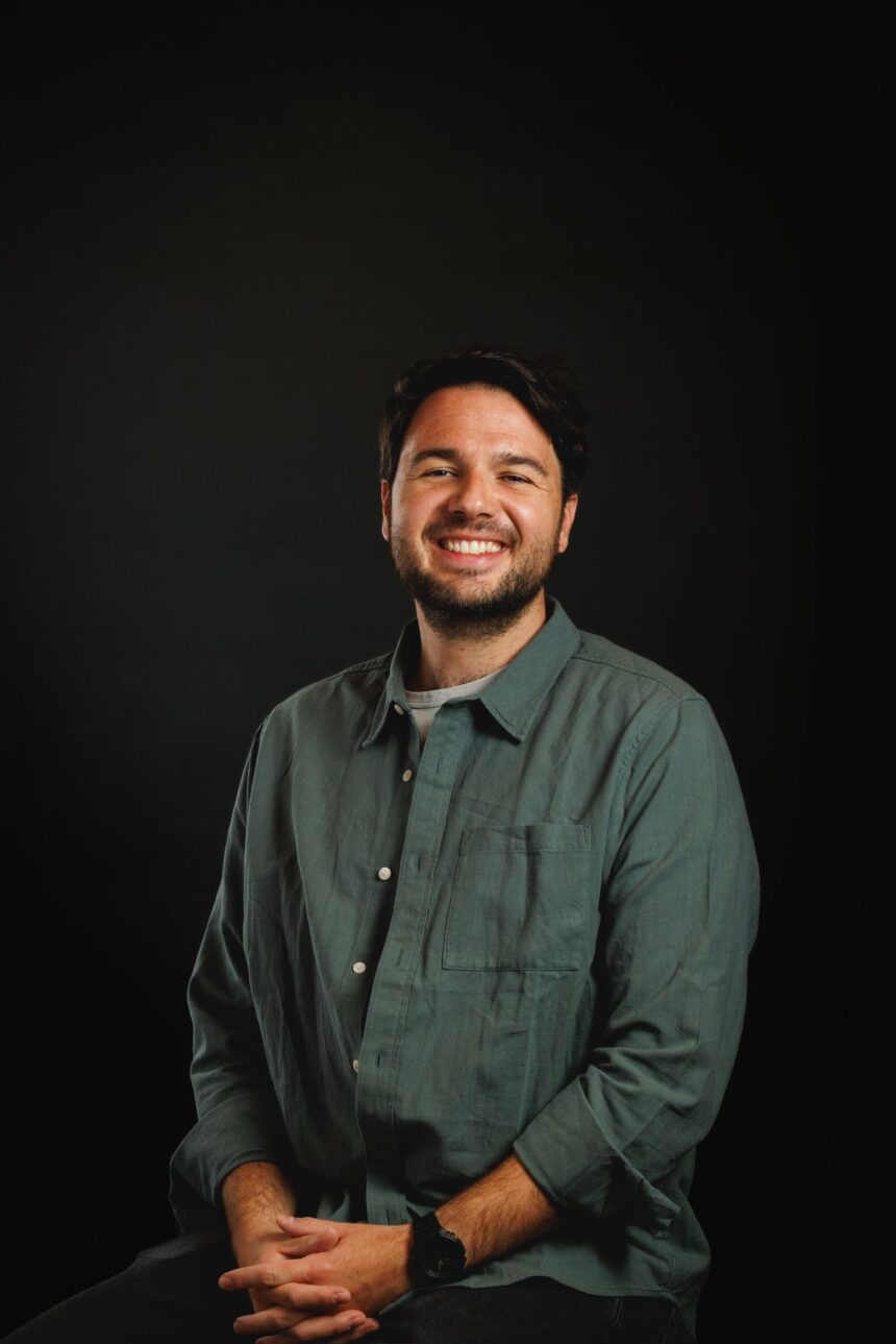 A smiling man with a beard wearing a green button-down shirt against a black background.
