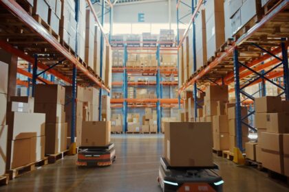 Two autonomous robots carrying boxes through a large, high-racked warehouse aisle.