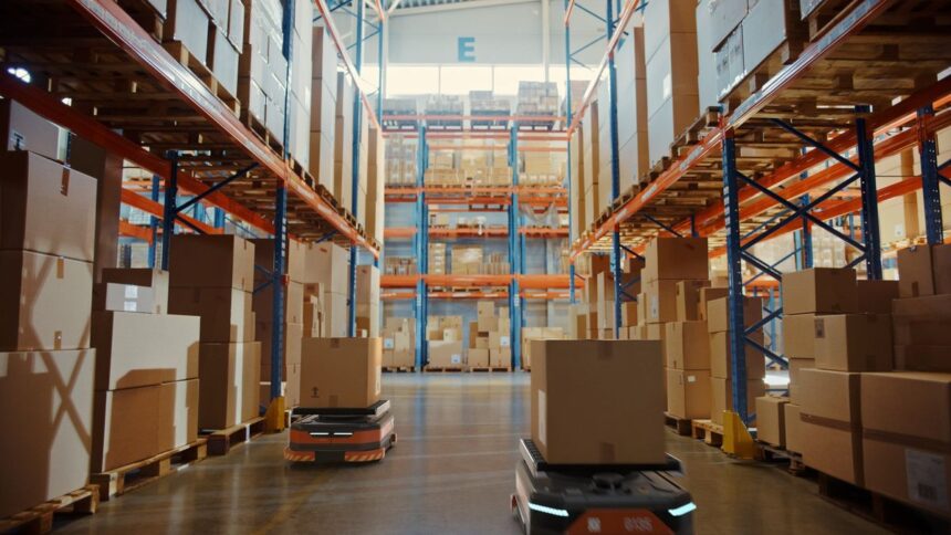 Two autonomous robots carrying boxes through a large, high-racked warehouse aisle.