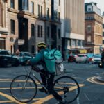 A Deliveroo cyclist wearing a yellow helmet rides through a city street intersection.