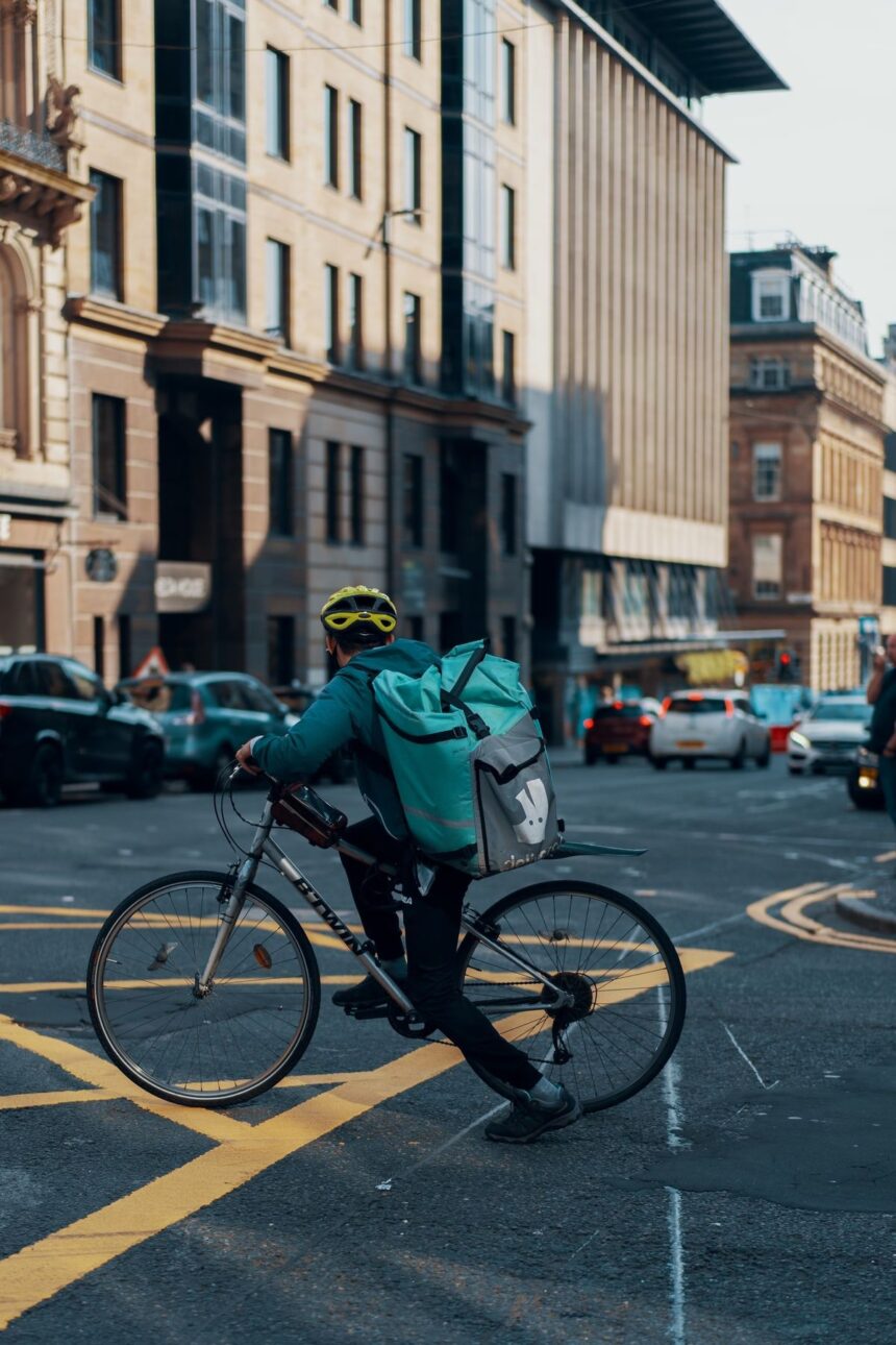 A Deliveroo cyclist wearing a yellow helmet rides through a city street intersection.