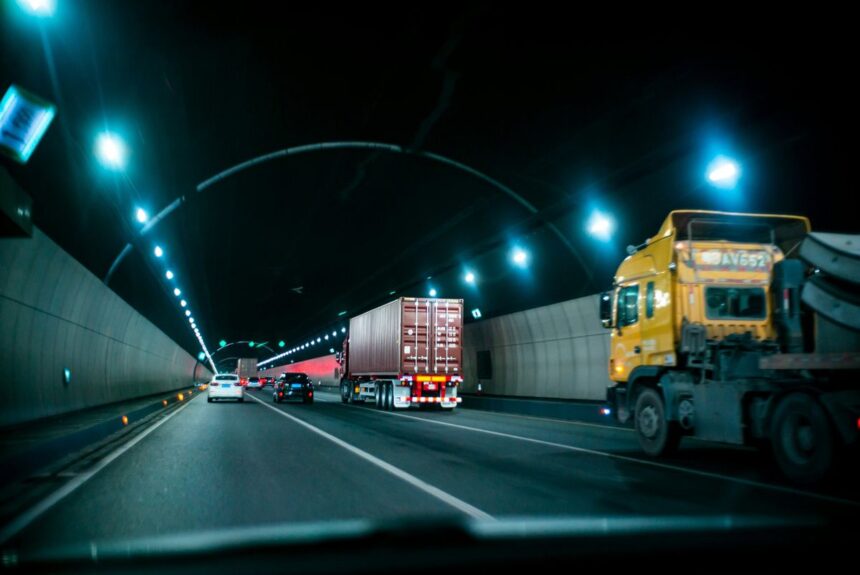 Cars and a semi-truck driving through a dimly lit highway tunnel at night.