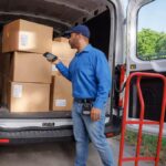 Delivery driver with a handheld device stands by a van filled with cardboard boxes.