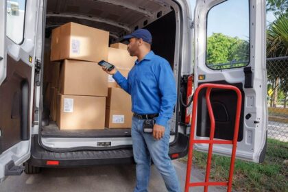 Delivery driver with a handheld device stands by a van filled with cardboard boxes.