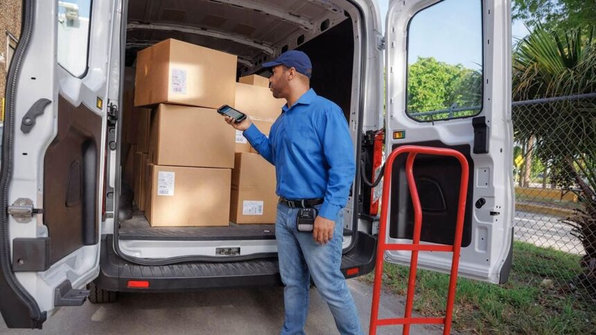 Delivery driver with a handheld device stands by a van filled with cardboard boxes.