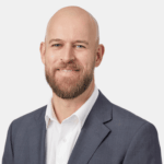 A smiling, bald man with a beard, wearing a gray suit and white shirt.