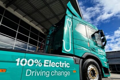 A teal Toll 100% electric semi-truck parked outside a building under a blue sky.
