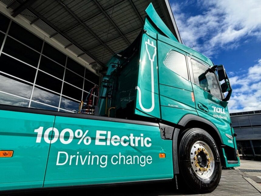 A teal Toll 100% electric semi-truck parked outside a building under a blue sky.