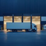 A semi-truck parked in front of a brightly lit industrial warehouse loading bay.