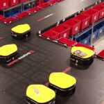 Yellow warehouse robots move packages past red and blue sorting bins on a track.