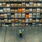 Worker using a tablet in a large warehouse with rows of inventory shelves.