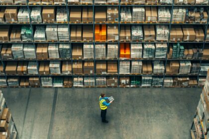 Worker using a tablet in a large warehouse with rows of inventory shelves.