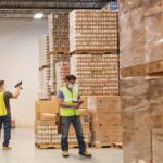 Warehouse workers wearing masks and vests scan products in a large, stocked storage facility.
