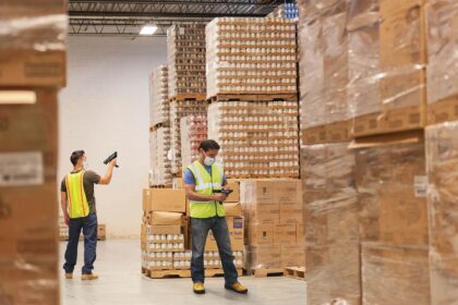 Warehouse workers wearing masks and vests scan products in a large, stocked storage facility.