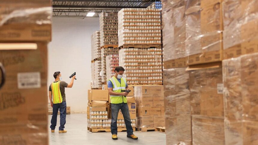 Warehouse workers wearing masks and vests scan products in a large, stocked storage facility.