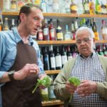 A shopkeeper and an older customer examining small bunches of leafy green produce.