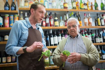 A shopkeeper and an older customer examining small bunches of leafy green produce.