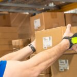 A worker wearing a wearable scanner lifts a cardboard box in a warehouse.