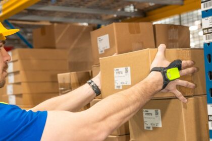 A worker wearing a wearable scanner lifts a cardboard box in a warehouse.