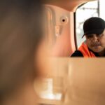 A delivery worker in an orange vest hands a cardboard box to a customer.