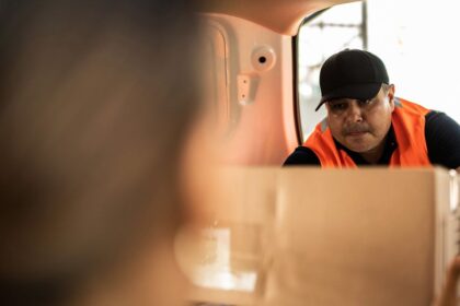 A delivery worker in an orange vest hands a cardboard box to a customer.