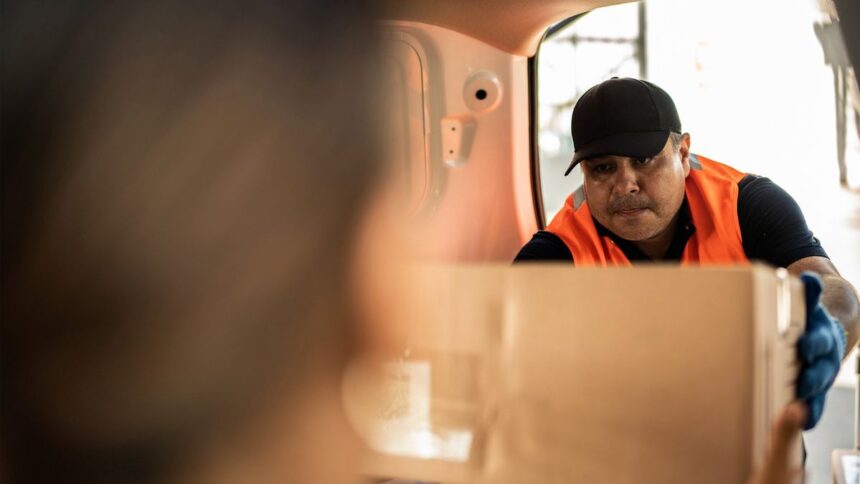 A delivery worker in an orange vest hands a cardboard box to a customer.