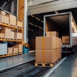 Warehouse loading dock with cardboard boxes on pallets and a semi-truck trailer being loaded.