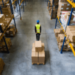 A warehouse worker pulls a pallet of cardboard boxes between rows of storage shelves.