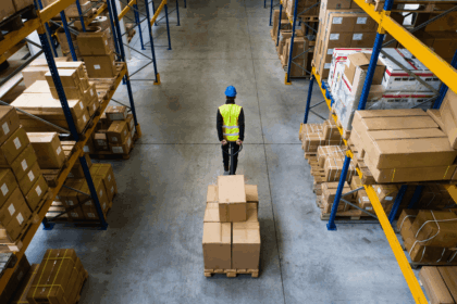 A warehouse worker pulls a pallet of cardboard boxes between rows of storage shelves.