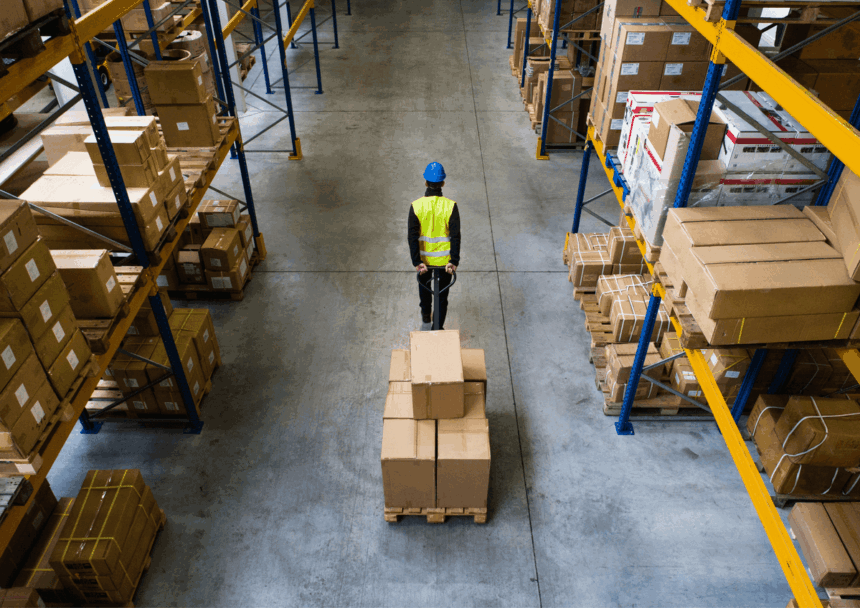 A warehouse worker pulls a pallet of cardboard boxes between rows of storage shelves.