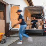 A worker uses a pallet jack while another organizes boxes inside a delivery van.