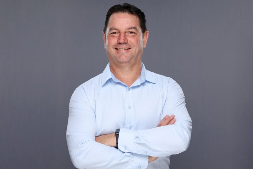 A middle-aged man in a light blue button-down shirt smiling with arms crossed.