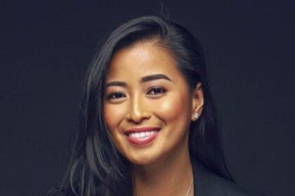 A smiling woman with long dark hair wearing a black blazer against a dark background.