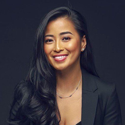 A smiling woman with long dark hair wearing a black blazer against a dark background.