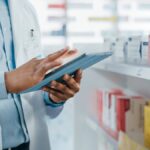 A pharmacist in a white coat uses a tablet in a pharmacy.