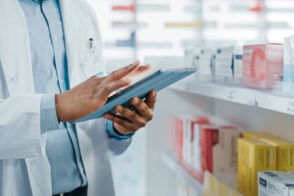 A pharmacist in a white coat uses a tablet in a pharmacy.