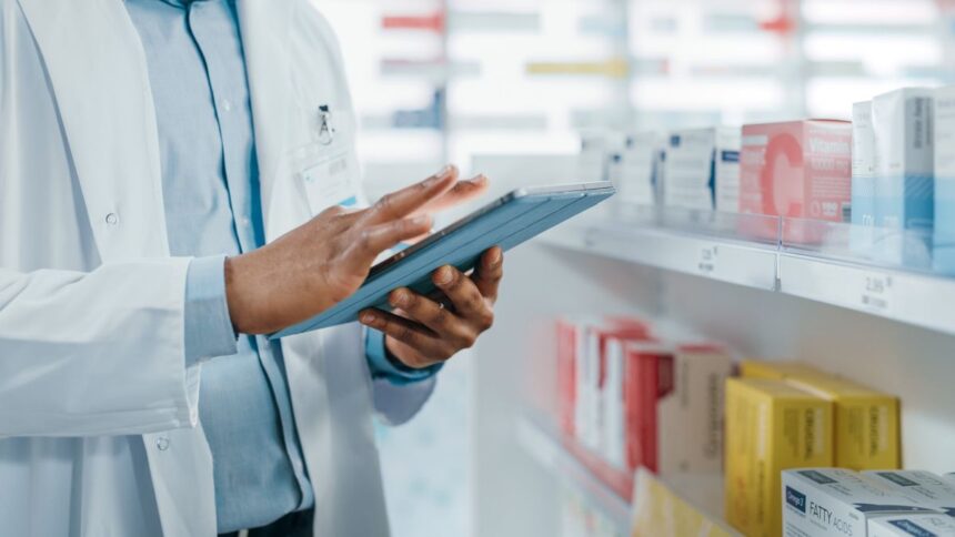 A pharmacist in a white coat uses a tablet in a pharmacy.